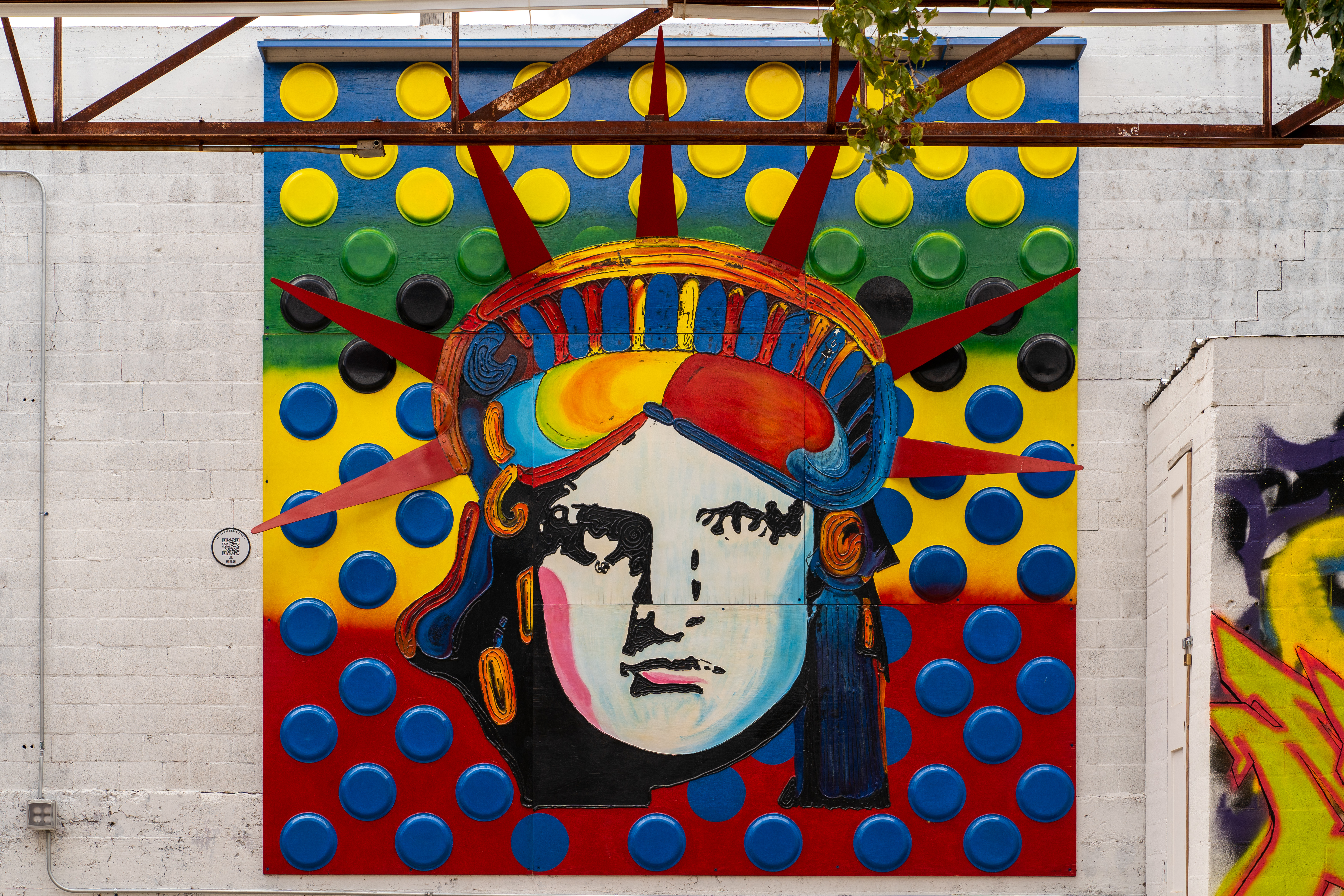 Abstract mural of the face of the Statue of Liberty in blue, green, yellow, and red in front of a white brick wall in a outdoor courtyard.