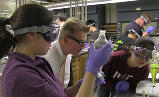Two students and Prof. John Dolhun lean over a chemistry lab bench to peer at a glass container filled with a neon green liquid. 